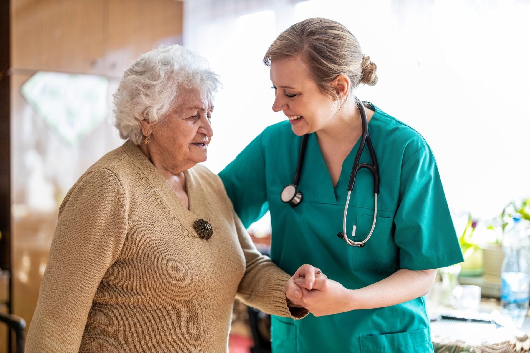 Nurse in scrubs smiling as she helps an older woman walk at home