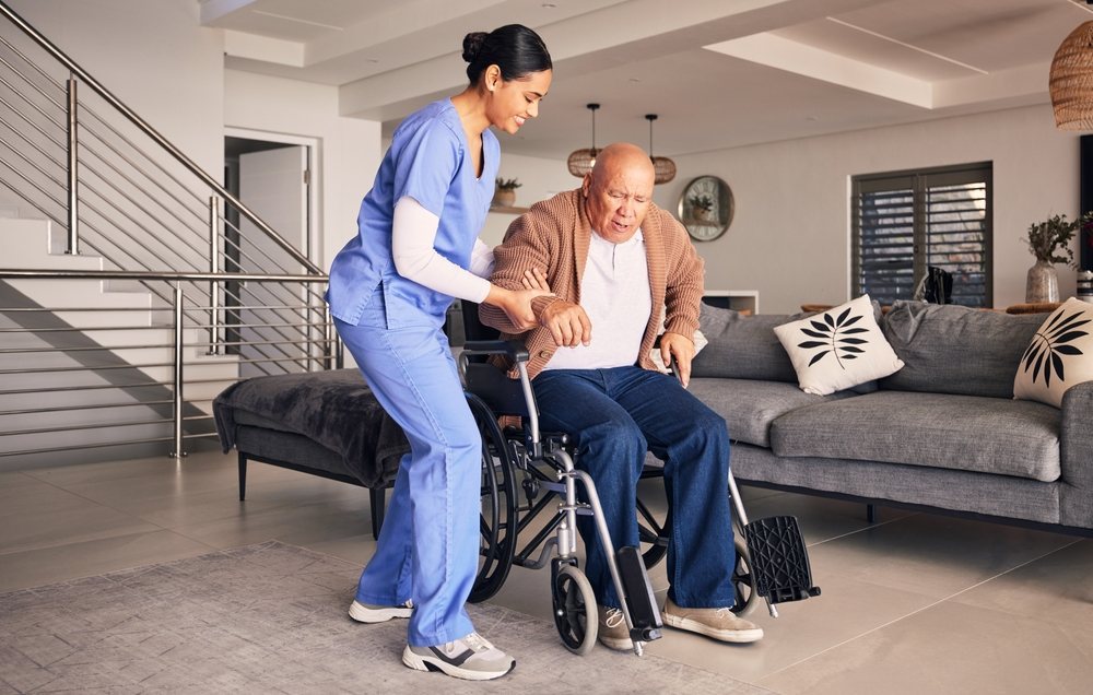 Nurse helping an older man rise from his wheelchair in his living room